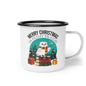 Könnte beinhalten: Weißer Emaillebecher mit schwarzem Rand und Henkel. Der Becher hat ein skurriles Design mit einer Eule, die einen Schal trägt und einen Zauberstab hält, mit dem Text "MERRY CHRISTMAS YA FILTHY MUGGLE" in einer festlichen Schriftart. Das Design umfasst Bücher, Geschenke und ein Schloss.