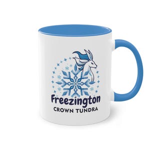 Op de afbeelding: Witte keramische mok met een blauwe binnenkant en handvat. De mok heeft een blauw en wit sneeuwvlokontwerp met een gestileerd wezen en de tekst "Freezington Crown Tundra" in blauw en zwart.