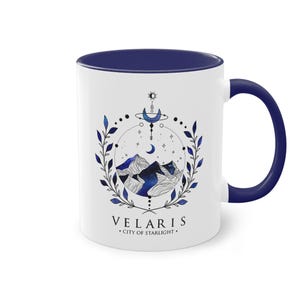 Könnte beinhalten: Weißer Keramikbecher mit marineblauem Innenraum und Griff. Der Becher hat ein kreisförmiges Design mit Bergen, einem Mond und dem Text "VELARIS - CITY OF STARLIGHT" in einer stilisierten Schriftart. Blaue Blätter und Punkte umgeben das Design.