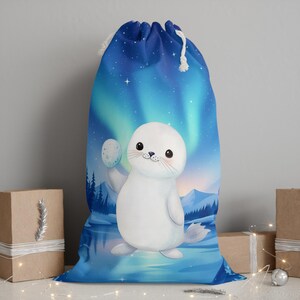 Könnte beinhalten: Blauer Geschenkbeutel mit Kordelzug, der einen Cartoon-Robben mit einem Mond zeigt. Der Hintergrund zeigt eine Winterlandschaft mit Nordlichtern. Der Beutel hat einen weißen Kordelzug.