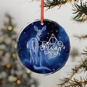 May include: A round, clear glass ornament with a blue watercolor background and a white deer illustration. The ornament features the text "Always" in elegant script, along with a symbol. A red ribbon is attached for hanging.