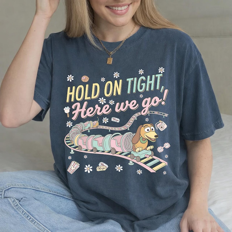 May include: Navy blue t-shirt with a colorful graphic featuring a cartoon dachshund on a roller coaster with the text "HOLD ON TIGHT, Here we go!". The design includes pastel colors and floral accents.