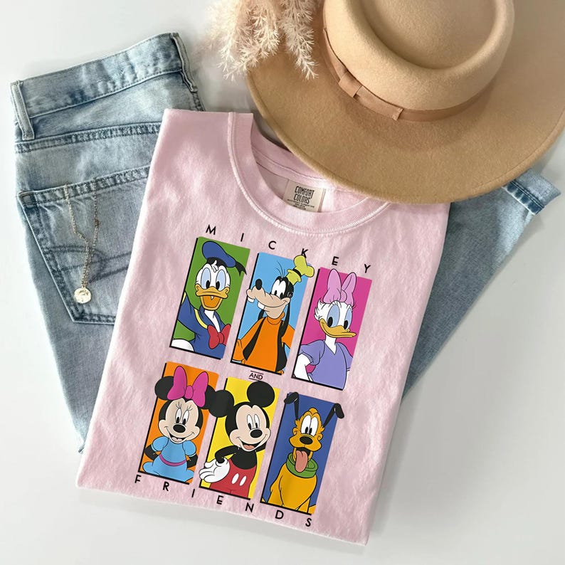 May include: A light pink t-shirt featuring the text "Mickey and Friends" with cartoon images of Mickey Mouse, Minnie Mouse, Donald Duck, Goofy, Daisy Duck, and Pluto. The shirt is paired with light blue jeans and a tan hat.