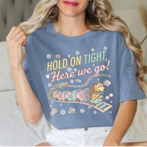 May include: Blue t-shirt with the text "HOLD ON TIGHT, Here we go!" and a cartoon image of Slinky Dog on a roller coaster. The design includes pastel colors and floral accents, creating a playful and whimsical aesthetic.