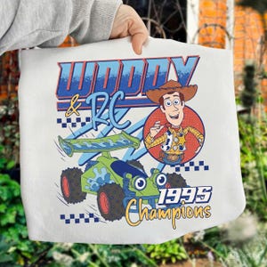 May include: White tote bag featuring a cartoon graphic of Woody and RC from the movie Toy Story. The design includes the text "Woody & RC" in blue and red, a green and blue race car, and the words "1995 Champions".