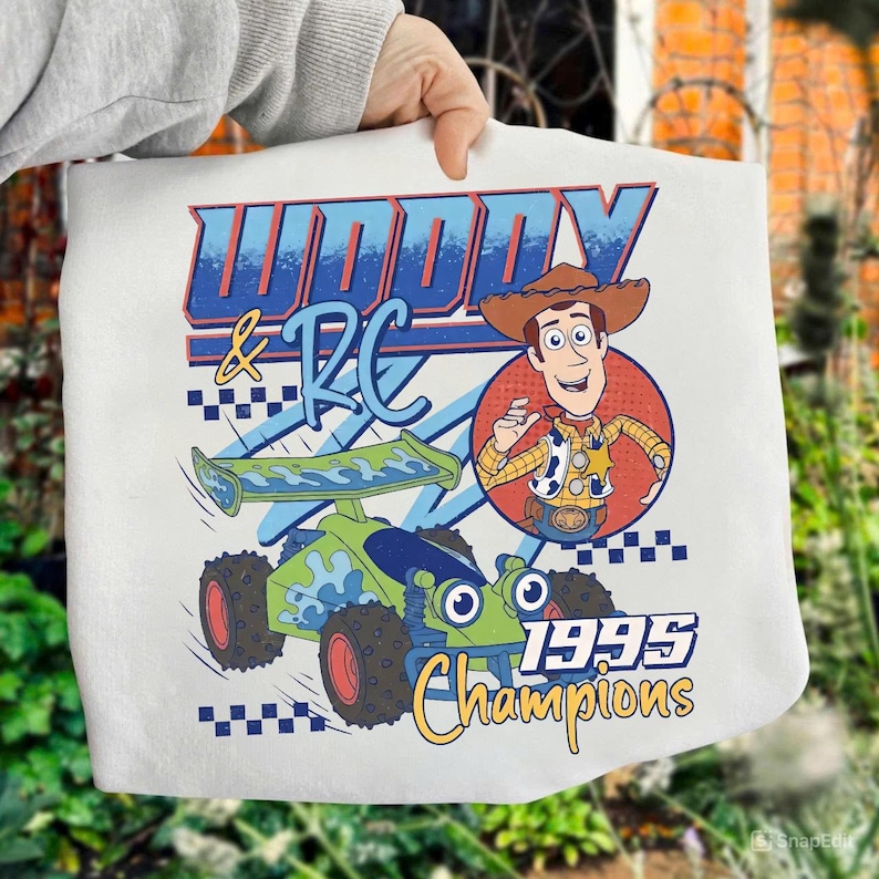 May include: White tote bag featuring a cartoon graphic of Woody and RC from the movie Toy Story. The design includes the text "Woody & RC" in blue and red, a green and blue race car, and the words "1995 Champions".