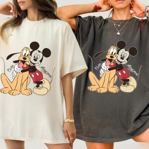 May include: Two oversized t-shirts, one cream and one charcoal, each featuring a cartoon graphic of Mickey Mouse hugging Pluto. The graphic includes the word "Pluto" and "Mickey Mouse" in script. The shirts have short sleeves and a relaxed fit.