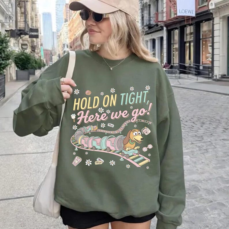 May include: Olive green sweatshirt with the text "HOLD ON TIGHT, Here we go!" and a cartoon image of a dachshund in a toy train. The design includes pastel colors and floral accents. The sweatshirt is a casual style.
