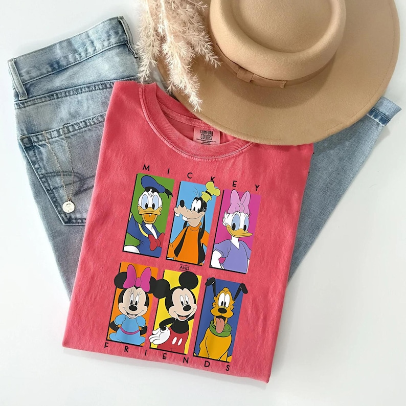 May include: A coral-colored t-shirt featuring a graphic of Mickey Mouse and friends. The shirt is paired with light-wash denim jeans and a tan fedora hat. The graphic includes Donald Duck, Goofy, Daisy Duck, Minnie Mouse, Mickey Mouse, and Pluto.