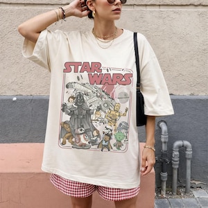 Könnte beinhalten: Cremefarbenes Star Wars T-Shirt mit Darth Vader, C-3PO, R2-D2 und anderen Charakteren. Das Design zeigt den Schriftzug "STAR WARS" in Rot und Weiß im Retro-Stil. Das T-Shirt ist oversized.