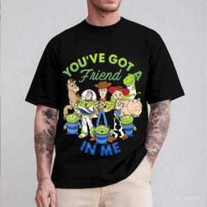 May include: Black t-shirt featuring a colorful graphic of characters from the animated film "Toy Story." The design includes Woody, Buzz Lightyear, Jessie, Rex, Hamm, and the Little Green Men, with the text "YOU'VE GOT A FRIEND IN ME."