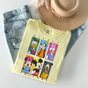 May include: A light yellow t-shirt featuring a graphic of Mickey Mouse and friends, including Donald Duck, Goofy, and Daisy Duck. The shirt is paired with light blue jeans and a beige hat. The text "MICKEY AND FRIENDS" is displayed on the shirt.