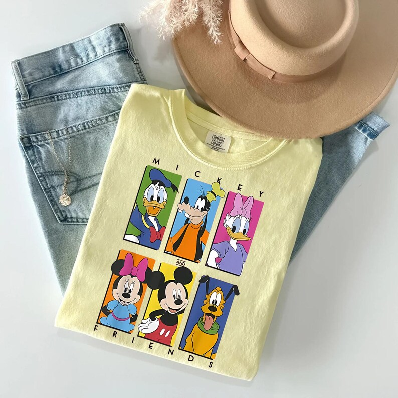 May include: A light yellow t-shirt featuring a graphic of Mickey Mouse and friends, including Donald Duck, Goofy, and Daisy Duck. The shirt is paired with light blue jeans and a beige hat. The text "MICKEY AND FRIENDS" is displayed on the shirt.