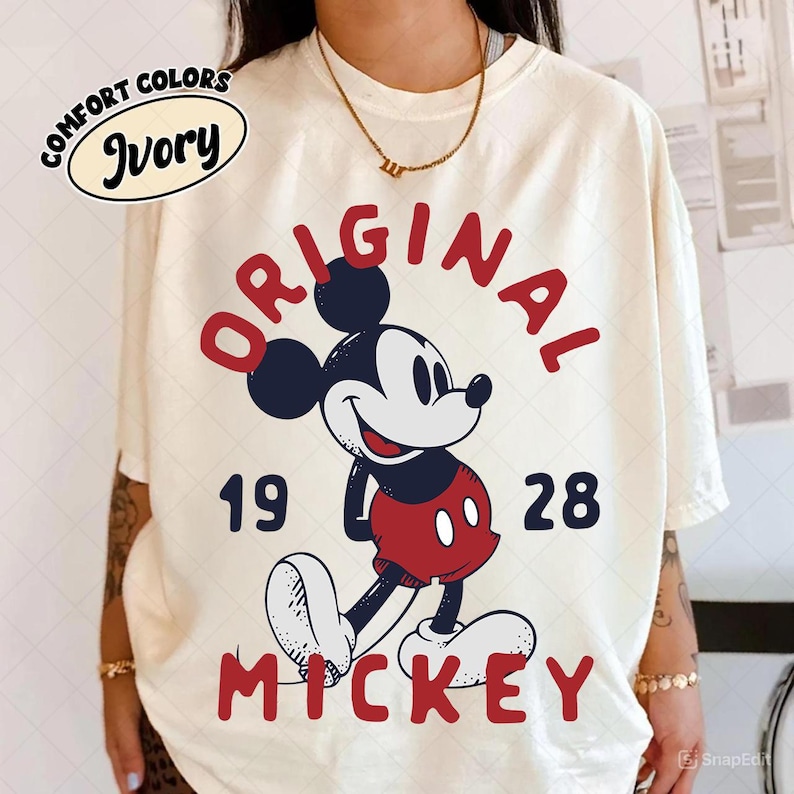 May include: Ivory-colored t-shirt featuring a classic Mickey Mouse graphic. The design includes the words "ORIGINAL MICKEY 1928" in red, with Mickey Mouse in his iconic red shorts and white gloves. The shirt also has the words "COMFORT COLORS" and "Ivory" in a circle.