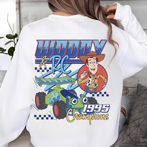 May include: White sweatshirt featuring a graphic of Woody and RC from Toy Story. The design includes a cartoon image of Woody in a cowboy hat, a green and blue race car, and the text "Woody & RC 1995 Champions."