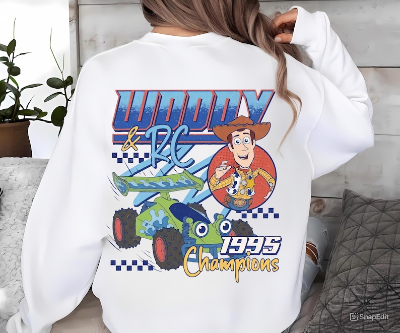 May include: White sweatshirt featuring a graphic of Woody and RC from Toy Story. The design includes a cartoon image of Woody in a cowboy hat, a green and blue race car, and the text "Woody & RC 1995 Champions."