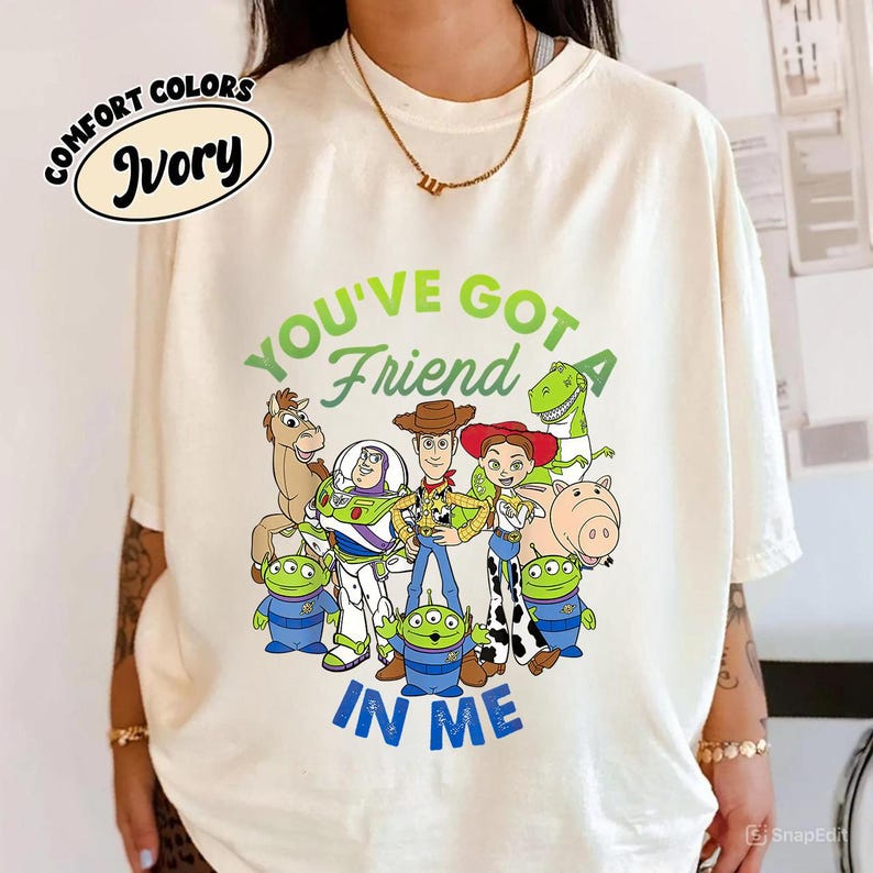 May include: Ivory-colored t-shirt featuring a graphic of characters from the animated film "Toy Story." The design includes Woody, Buzz Lightyear, Jessie, and other characters, with the text "YOU'VE GOT A FRIEND IN ME" in green and blue.