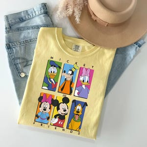 May include: A light yellow t-shirt featuring the text "Mickey and Friends" with cartoon images of Mickey Mouse, Minnie Mouse, Donald Duck, Daisy Duck, Goofy, and Pluto. The shirt is laid on top of blue jeans and a tan hat.