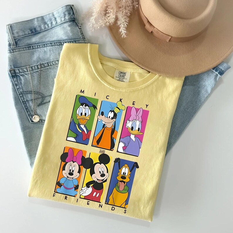 May include: A light yellow t-shirt featuring the text "Mickey and Friends" with cartoon images of Mickey Mouse, Minnie Mouse, Donald Duck, Daisy Duck, Goofy, and Pluto. The shirt is laid on top of blue jeans and a tan hat.