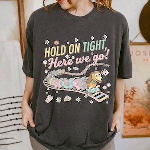 May include: Dark gray t-shirt featuring a colorful graphic of a dachshund riding a roller coaster with the text "HOLD ON TIGHT, Here we go!". The design includes pastel colors and floral accents, creating a playful and whimsical aesthetic.