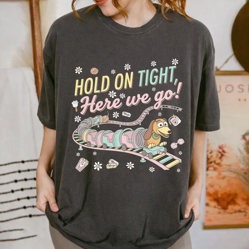 May include: Dark gray t-shirt featuring a colorful graphic of a dachshund riding a roller coaster with the text "HOLD ON TIGHT, Here we go!". The design includes pastel colors and floral accents, creating a playful and whimsical aesthetic.