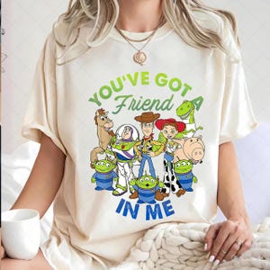 May include: Cream-colored t-shirt featuring a graphic of characters from the animated film Toy Story. The text "YOU'VE GOT A FRIEND IN ME" is printed in green and blue. The design includes Woody, Buzz Lightyear, and other characters.