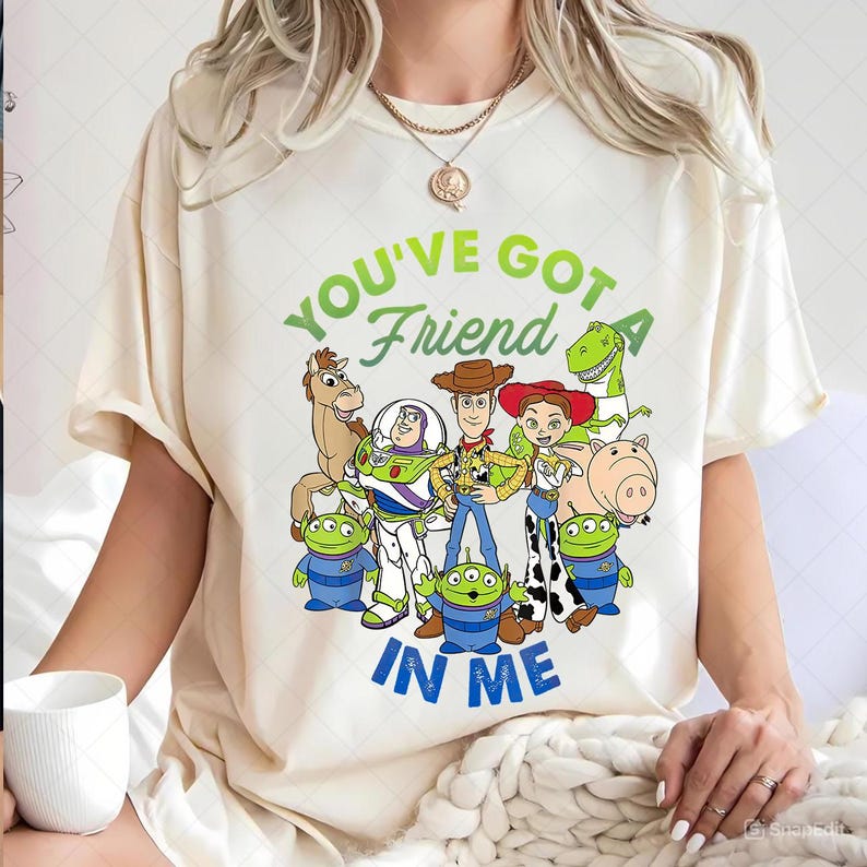 May include: Cream-colored t-shirt featuring a graphic of characters from the animated film Toy Story. The text "YOU'VE GOT A FRIEND IN ME" is printed in green and blue. The design includes Woody, Buzz Lightyear, and other characters.