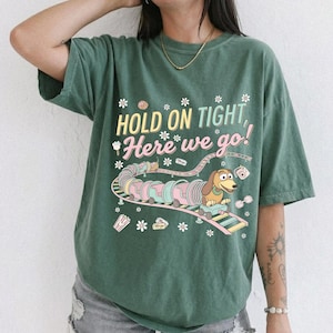 May include: Green t-shirt with the phrase "HOLD ON TIGHT, Here we go!" and a cartoon image of a dachshund dog riding a roller coaster. The design includes pastel colors and floral accents.