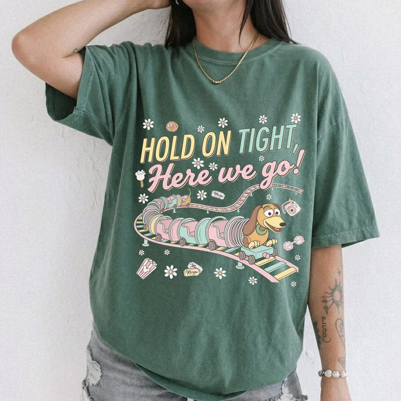 May include: Green t-shirt with the phrase "HOLD ON TIGHT, Here we go!" and a cartoon image of a dachshund dog riding a roller coaster. The design includes pastel colors and floral accents.