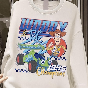 以下が含まれることがあります： トイ・ストーリーのウッディとRCのグラフィックが特徴のクリーム色のスウェットシャツ。「Woody & RC 1995 Champions」の文字と、ウッディの漫画の画像、緑と青のRCカーがデザインされています。