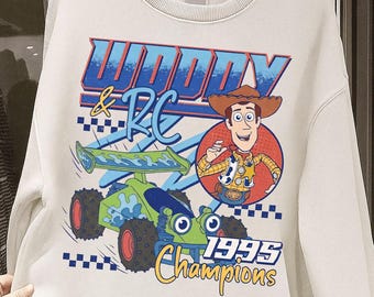 Puede incluir: Sudadera color crema con un gráfico de Woody y RC de Toy Story. El diseño incluye el texto "Woody & RC 1995 Champions" con una imagen de dibujos animados de Woody y un coche RC verde y azul.