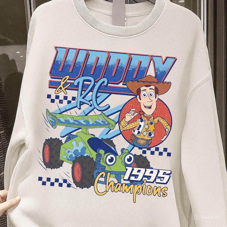 May include: Cream-colored sweatshirt featuring a graphic of Woody and RC from Toy Story. The design includes the text "Woody & RC 1995 Champions" with a cartoon image of Woody and a green and blue RC car.