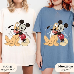 May include: Ivory and blue jean t-shirts featuring a graphic of Mickey Mouse hugging Pluto. The cartoon characters are in a warm embrace, with Pluto's tongue sticking out. The text "Mickey Mouse" is printed on the shirts.