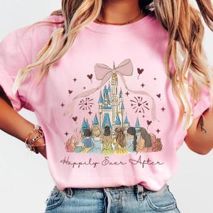 May include: Pink crew neck t-shirt featuring a castle illustration with a large bow and the text "Happily Ever After." The graphic shows several figures in front of the castle, with hearts and stars scattered around the design.