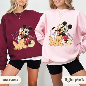May include: Maroon and light pink sweatshirts featuring a graphic of Mickey Mouse hugging Pluto. The cartoon characters are in a warm embrace. The sweatshirts have long sleeves and a crew neck. The text "Mickey Mouse" and "Pluto" are also visible.