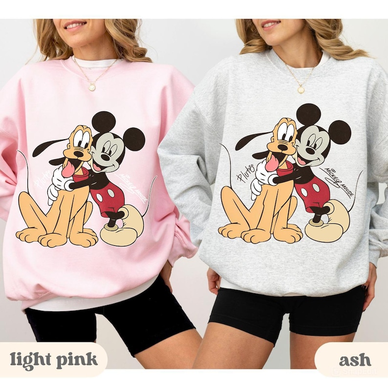 May include: Two sweatshirts, one light pink and one ash gray, each featuring a cartoon graphic of Mickey Mouse hugging Pluto. The text "Mickey Mouse" and "Pluto" are visible. The sweatshirts are shown on a person.