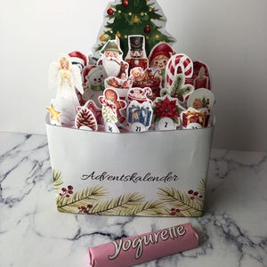 以下が含まれることがあります： クリスマスツリーと華やかなフィギュアが描かれた白いアドベントカレンダー。星、サンタクロース、くるみ割り人形、その他お祝いをテーマにしたイラストが特徴です。「Adventskalender」の文字が前面に印刷されています。ピンク色のチョコレートバーが前景にあります。