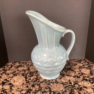 May include: A light blue ceramic pitcher with a handle. The pitcher has a textured, diamond-patterned lower half and a fluted upper portion. The pitcher is approximately 10 inches tall.