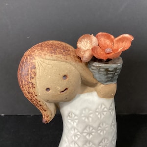 May include: A ceramic figurine of a girl holding a small basket with orange flowers. The girl has brown hair and a smiling face. Her white dress has a starburst pattern. The figurine is a decorative item.