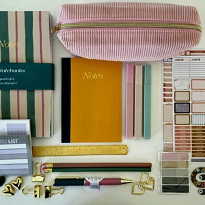 May include: A stationery set including a striped A5 notebook pack, a mustard yellow notebook, a pink corduroy pencil case, three pens, a ruler, pencils, a pen, sticker sheets, sticky notes, heart-shaped paper clips, and gold binder clips.