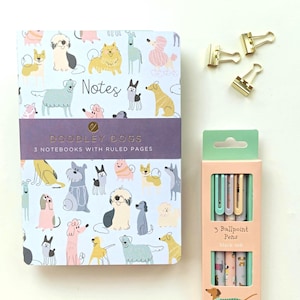 Könnte beinhalten: Ein Notizbuch mit Hundemuster, dem Text "Notes" und "DOODLEY DOGS 3 NOTEBOOKS WITH RULED PAGES", eine Schachtel mit drei Kugelschreibern mit schwarzer Tinte und drei goldfarbene Metall-Binderclips. Die Stifteschachtel zeigt eine Dackel-Illustration.