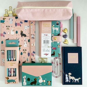 Puede incluir: Un conjunto de papelería que incluye un estuche de pana rosa, un cuaderno con estampado de perros, una agenda 2027, una caja de tres bolígrafos, clips dorados, dijes en forma de corazón y un clip con diseño de perro.