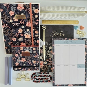 Op de afbeelding: Een briefpapierset met een bloemrijk notitieboek met het woord NOTES, een kleiner bijpassend notitieboek, een weekplanner, een etui, een gouden liniaal, een gouden pen met een libel, twee paarse stiften, drie gouden klemmen en paperclips.