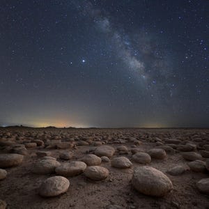 May include: A night scene showing the Milky Way and a shooting star streaking across the sky. The foreground is a field of rounded stones. The sky is dark blue, filled with stars, and the Milky Way is a bright band.