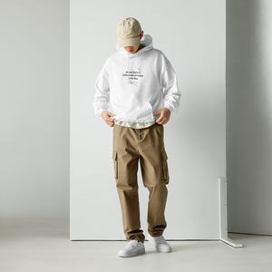 Peut inclure: Un sweat à capuche blanc avec une poche avant et une capuche à cordon. Le sweat-shirt porte du texte sur le devant. La personne porte un pantalon cargo kaki, une casquette de baseball beige et des baskets blanches.