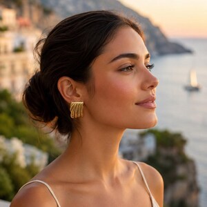 May include: A gold-toned earring featuring a series of five parallel, curved, vertical bars that create a fan-like, ribbed texture. The earring has a polished metallic finish and a rectangular shape that sits flush against the earlobe.