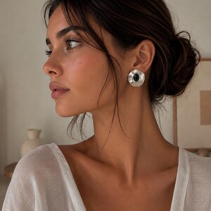 May include: A circular stud earring featuring a hammered silver-tone metal finish with a smooth, oval-shaped black stone set in the centre. The earring has a concave, wavy edge design that catches the light, creating a textured appearance.