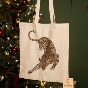 Könnte beinhalten: Beige Tragetasche mit einer detaillierten Illustration eines Leoparden. Der Leopard hat ein geflecktes Fell und ist in einer dynamischen Pose dargestellt. Die Tasche hat lange Henkel und den Text "blablabag".