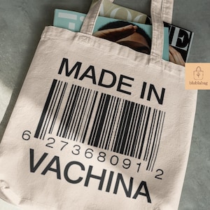 Può includere: Una borsa tote beige con la scritta "MADE IN VACHINA" e un design a codice a barre nero. La borsa contiene riviste e ha un piccolo logo sul lato. La borsa ha un design semplice ed è realizzata in tessuto chiaro.