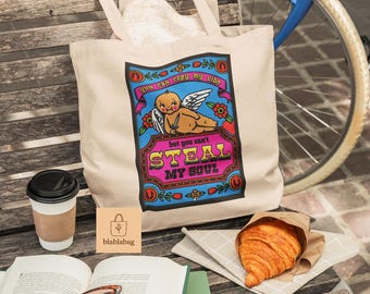 May include: A beige tote bag with a colourful graphic that reads "You can copy my vibe but you can't steal my soul." The bag is on a wooden bench with a coffee cup, book, glasses, and a croissant. A bicycle is in the background.